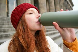 Portrait of redhead woman drinking from thermos, sitting on street stairs and enjoys hot drink from flask photo