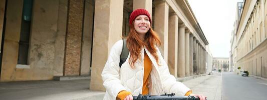 joven sonriente pelirrojo chica, estudiante paseos eléctrico scooter, alquila eso y viajes alrededor ciudad foto
