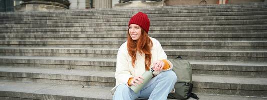 Young redhead female tourist rests during her trip, opens thermos and drinks hot tea, having a break after sightseeing photo