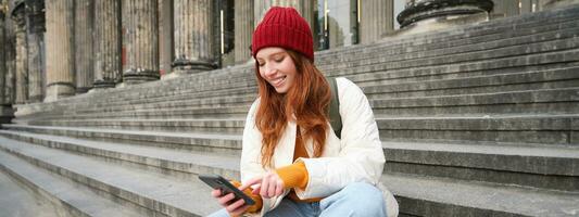 Portrait of young urban girl in red hat, sits on stairs near museum, holds mobile phone, connects to public wifi and surfs net, uses smartphone apps photo