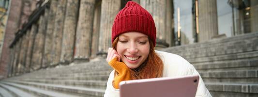 Portrait of young redhead woman sitting outdoors on stairs, reading ebook on digital tablet, wearing red hat and warm clothes photo