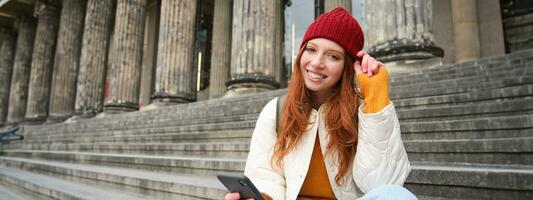 Portrait of young urban girl in red hat, sits on stairs near museum, holds mobile phone, connects to public wifi and surfs net, uses smartphone apps photo