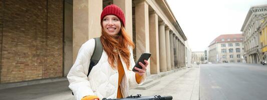 Redhead girl, tourist with backpack, uses mobile phone to rent escooter on streets of European city photo