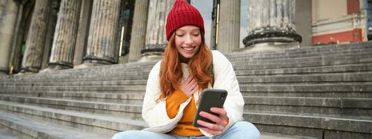 Young smiling woman with long red hair, sits near building on stairs with smartphone, using mobile application, sending message photo