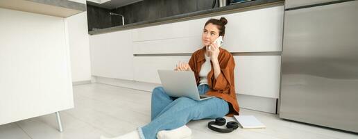 Young woman casually sits on floor with laptop, makes a phone call, talking on telephone photo