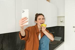 Happy woman using smartphone to take selfie, eating an apple on camera, records video for social media application, sitting on kitchen counter photo