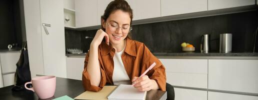 retrato de moderno joven mujer en anteojos, estudiante sentado a hogar y haciendo tarea, escritura abajo información en cuaderno y sonriente, trabajando en proyecto foto