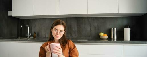 Portrait of young woman enjoying cup of coffee in peace, sitting at home, holding mug with herbal tea, wearing glasses, relaxing at in kitchen photo