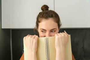 Cute woman hides face behind notebook, looking aside, smiling with eyes photo