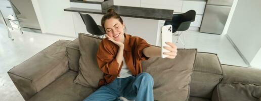 retrato de hermosa sonriente mujer, toma selfie a hogar, posando en sofá, sostiene teléfono inteligente con extendido mano foto