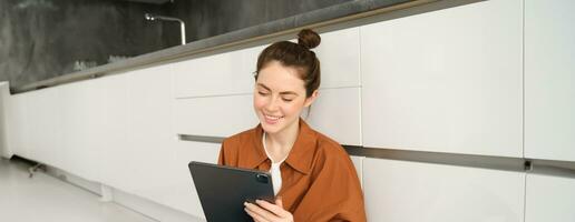 Portrait of young beautiful woman sitting on kitchen floor with digital tablet, browsing news feed, social media app on gadget, smiling and looking happy photo