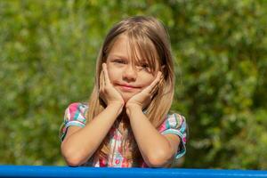 Portrait of caucasian little girl of 6 7 years smiling holding hands under chin and looking at camera over green trees photo