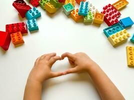 Child shows a heart against background of colorful building blocks on white background. Top view. photo