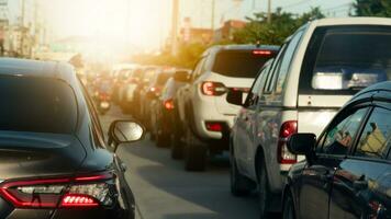 Rear view of the car and turn on brake light. Queues extend in a long line. On the asphalt road. illuminated by headlights. Urban traffic with small buildings ahead and green trees in evening. photo