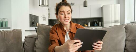 Portrait of young woman looking stressed and tensed at digital tablet, playing difficult level video game, sitting on couch, using gadget, spending time at home photo