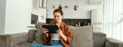 retrato de joven mujer sentado en sofá en vivo habitación, acecho películas en digital tableta, leyendo en dispositivo, disfrutando fin de semana a hogar foto