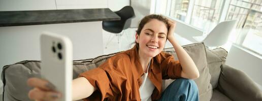 retrato de elegante, moderno joven mujer tomando selfie en teléfono inteligente, sentado en sofá con móvil teléfono, tomando imágenes para su estilo de vida Blog foto