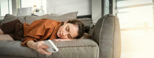 cerca arriba retrato de descansando mujer, acostado en sofá a hogar, leyendo Noticias en teléfono inteligente, desplazamiento social medios de comunicación aplicación en móvil teléfono y relajante en sofá foto