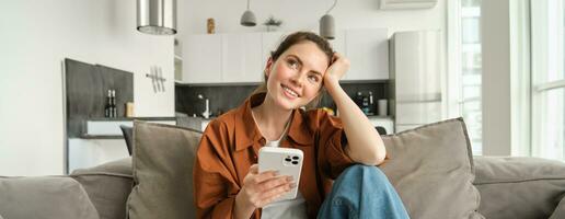 Smiling brunette woman sitting on sofa with smartphone, looking thoughtful, thinking while using mobile phone app, ordering on application, doing online shopping and resting at home photo