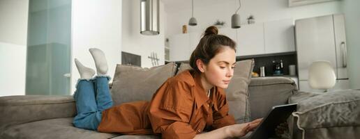 Weekend and lifestyle concept. Young woman lying on couch with digital tablet, scrolling social media, reading ebook or watching tv series on app photo