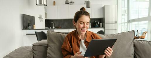 Portrait of young brunette woman reading on digital tablet, watching tv series on her application, sitting on couch in living room photo