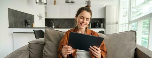 Portrait of young brunette woman reading on digital tablet, watching tv series on her application, sitting on couch in living room photo