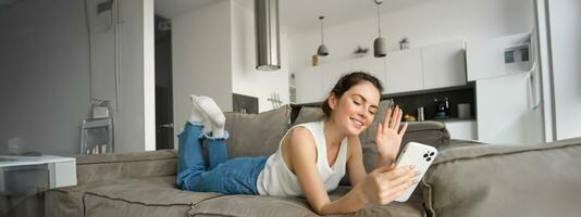 Happy cute female model, lying on couch, waving hand at smartphone, saying hi, connects to video call, resting at home and talking to friend photo