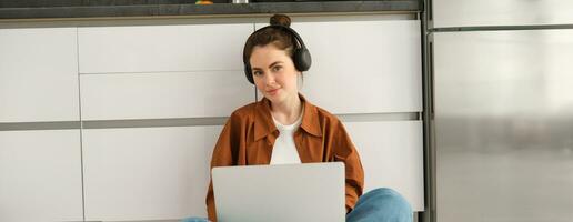 Young woman, student doing homework on laptop, sitting on floor, listening to music in headphones and working from home photo