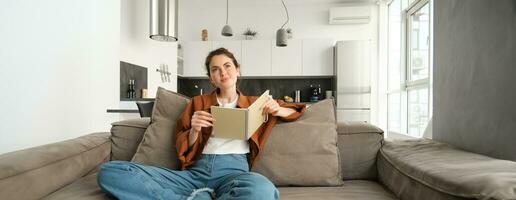 retrato de joven sonriente hermosa mujer, sentado en sofá en su vivo habitación, participación estudiar material, haciendo tarea, leyendo notas en cuaderno foto