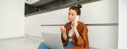 mujer mirando aburrido y aburrido mientras sentado en piso y escuchando a conversación, tiene ordenador portátil en vueltas, esperando en línea, haciendo teléfono llamada foto