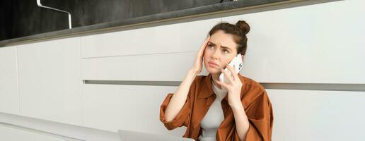 Portrait of woman calling her doctor on the phone, holding smartphone, frowning and touching her head, has painful headache or migraine, feeling unwell photo