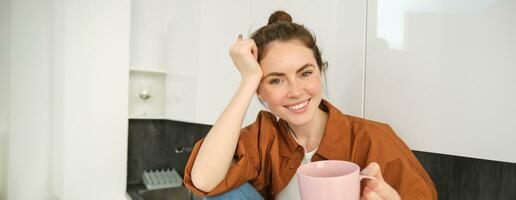 Portrait of woman with cup of coffee in hand, sits in kitchen and smiles at camera photo