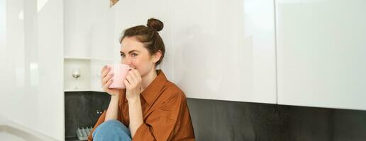 retrato de joven mujer con taza de café, se sienta en cocina y bebidas aromático bebida a hogar, sostiene té jarra foto