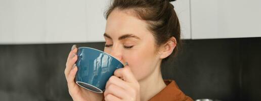 retrato de hermosa joven mujer, disfrutando delicioso aroma de recién hecho café, participación taza, en pie cerca máquina en el cocina, Bebiendo desde jarra foto