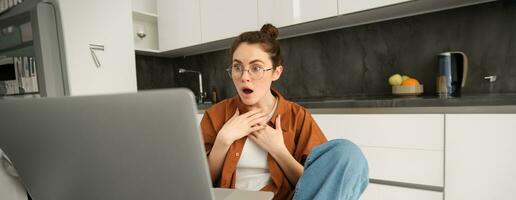 Portrait of woman looking at laptop with shocked face, seeing big news. Female student in kitchen, looking at her computer with amazement, gasping in surprise photo