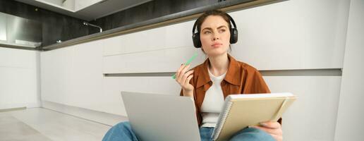 Portrait of young woman, modern student sits at home on floor and doing homework on laptop, listens music in headphones while making assignment or project for college photo