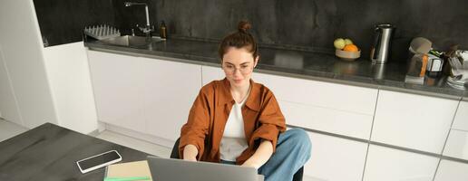 Portrait of beautiful young woman studying at home, sitting with laptop and documents in kitchen. Selfemployed business owner typing on computer, sending emails to clients photo