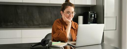 Portrait of young woman working from home, sets up workplace in kitchen, using laptop. Student doing homework on computer photo