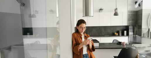Portrait of cute young woman standing at home with smartphone, reading message on mobile phone, using application, order online, paying utility bill in app photo