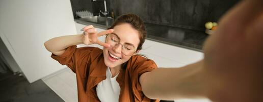 Portrait of carefree young woman in glasses, taking selfie from home, lifestyle blogger making photos in kitchen, laughing and smiling, using smartphone camera