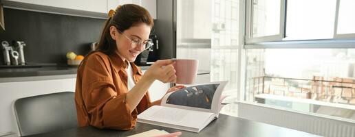 retrato de mujer leyendo a hogar, voltear paginas de favorito libro, relajante en cocina con taza de café, vistiendo lentes foto