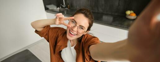 Selfie of happy young and carefree woman, taking photo on mobile phone with extended hand, posing and smiling, sitting in kitchen in glasses