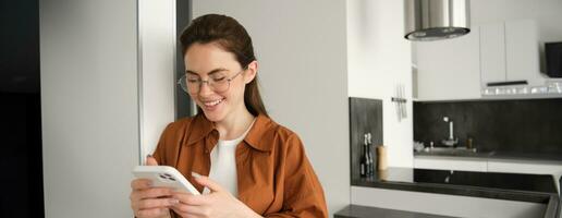 Portrait of cute young woman standing at home with smartphone, reading message on mobile phone, using application, order online, paying utility bill in app photo