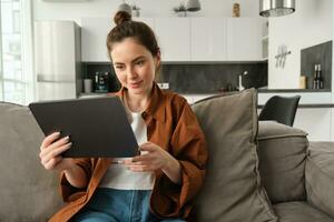 Portrait of smiling beautiful woman, sitting on couch, watching videos on digital tablet, reading ebook on her device, resting at home photo