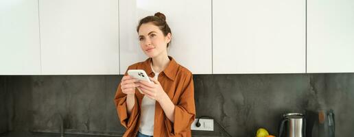 Image of young woman with smartphone, looking with thoughtful expression, buying online using mobile app, orders food delivery with phone application photo