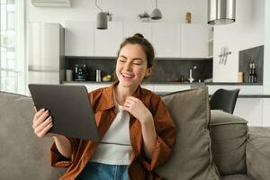 Portrait of woman laughing over funny video on digital tablet, watching movie on her gadget streaming service, sitting on sofa at home, reading ebook photo