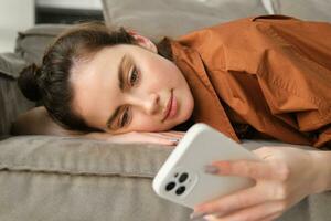 Close up portrait of smiling, beautiful young woman lying on sofa at home, checking messages on smartphone, resting on couch and looking at mobile phone photo