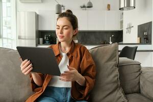 Portrait of female model reading on digital tablet ebook, watching tv series on her gadget application, sitting in living room at home photo