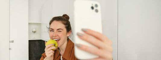 retrato de feliz, sonriente joven mujer registros sí misma, toma selfie mientras comiendo un manzana en el cocina, utilizando teléfono inteligente aplicación, hace fotos con móvil teléfono