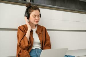 Young woman, student doing homework on laptop, sitting on floor, listening to music in headphones and working from home photo